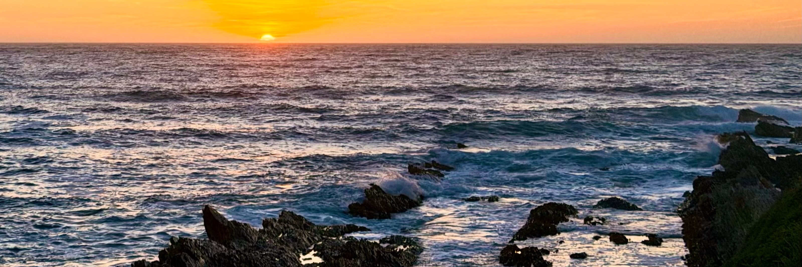 The sun half-set in an orange sky over silver and blue ocean waves. Waves crash over crags of rock in the foreground.