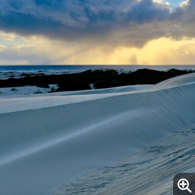 From over the ocean, an isolated rain cloud, backlit by the sun, approaches cool blue sand dunes.