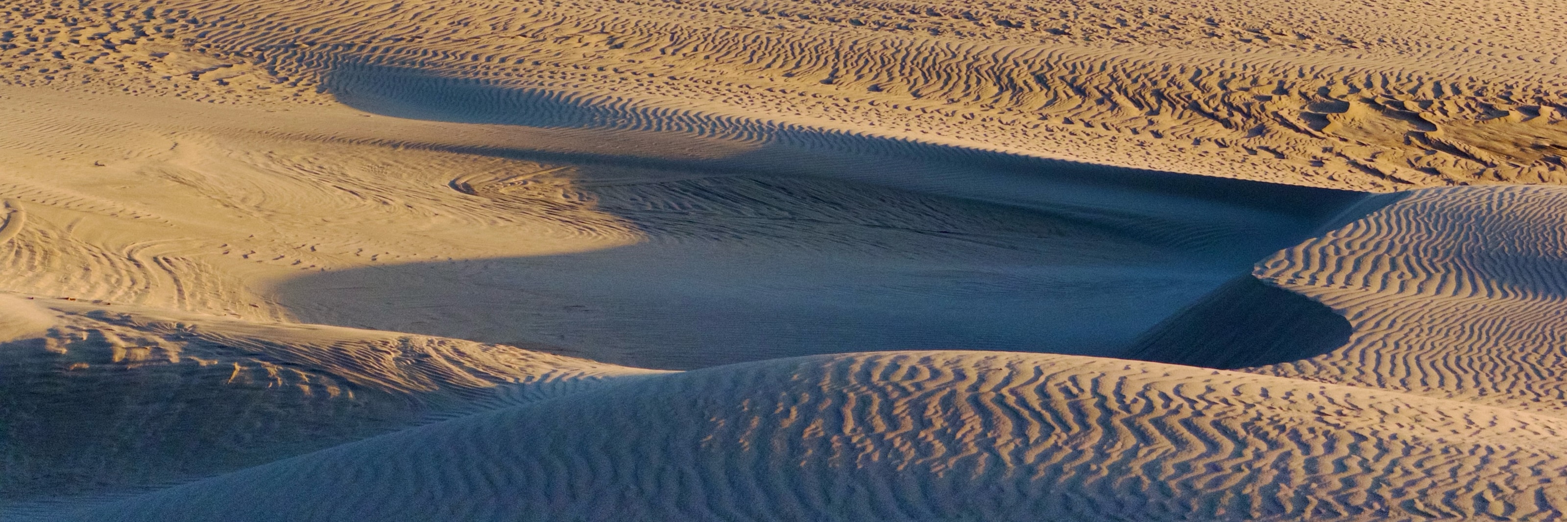 Ripples of alternating orange and blue zig-zag shapes along wind-blown sand dunes at sunset.
