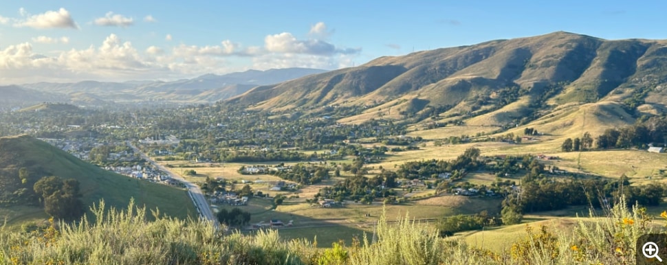 Rolling hills under a blue, partially cloudy sky. Smaller hills stand out of the valley to the left, with the outskirts of a town (San Luis Obispo) nestled in between.