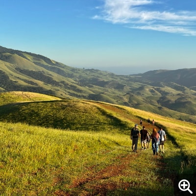 Nine hikers descend a rugged jeep road. Rolling green hills in the background give way to a hazy ridge. 